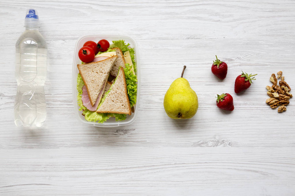 Healthy school lunch box with fresh organic vegetables sandwich, walnuts, fruits and bottle of water on white wooden background, flat lay. From above. Copy space. - Photo, Image