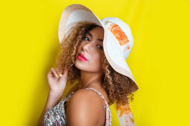 Beauty portrait of young african american girl with afro hairstyle in summer hat. Girl posing on yellow background, looking at camera. - Photo, image