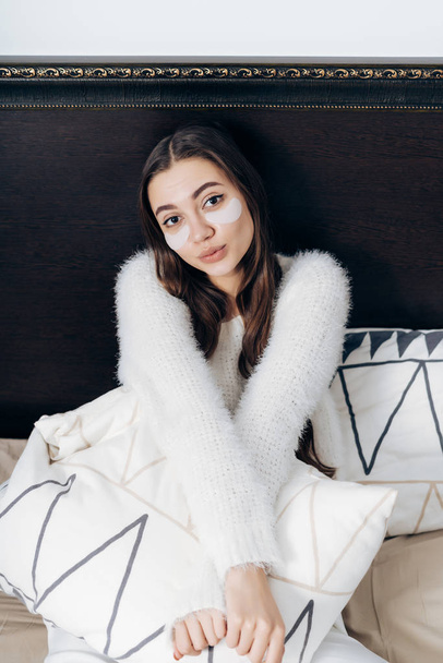 lovely long-haired girl in white pajamas woke up early in the morning, sits in bed, under the eyes white patches - Foto, afbeelding