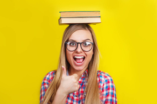 young woman in glasses holding books on her head. Smiling young stylish student is standing with books on yellow background - Photo, image