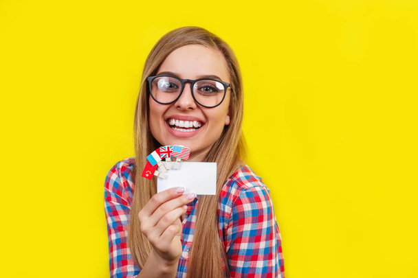 Young woman with card and flags of different speaking countries. Studio portrait of young beautiful female student on yellow background - Photo, image