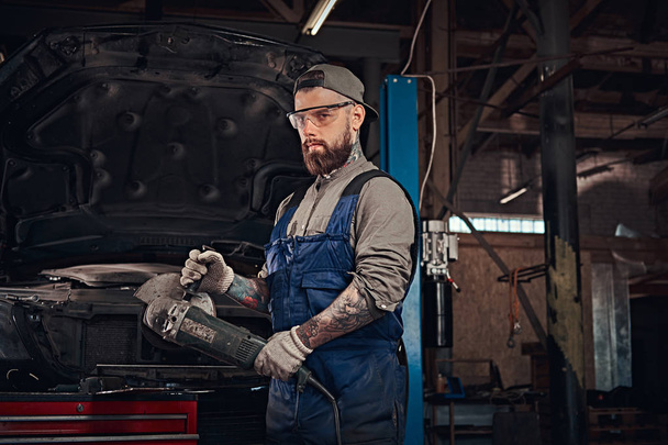 Brutal auto mechanic in a uniform and safety glasses working with an angle grinder while standing against a broken car in repair garage. - Foto, Imagen