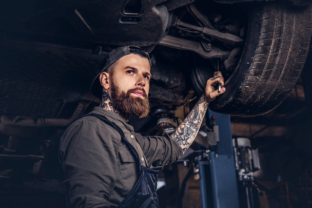 Bebaarde automonteur in een uniform herstellen de vering van de auto's met een moersleutel staand onder de opheffing van de auto in de garage voor reparatie. - Foto, afbeelding