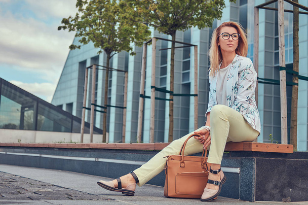 Όμορφη μόδας γυναίκα σε μοντέρνα ρούχα και τα γυαλιά με μια τσάντα, κάθεται σε ένα παγκάκι εναντίον ενός ουρανοξύστη. - Φωτογραφία, εικόνα