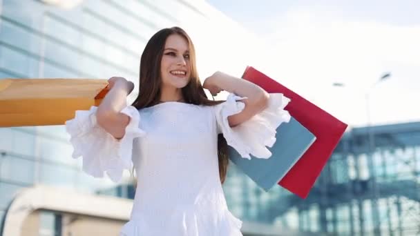 Pretty fashion model in white dress poses with shopping bags before a modern glass building - Metraje, vídeo