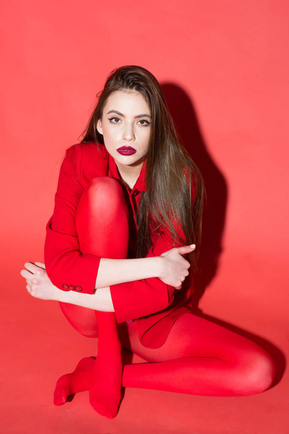 Young beautiful female model in red tights and jacket isolated on scarlet background. Portrait of beautiful girl with stylish make-up and burgundy lips sitting embracing her legs on floor. - Fotoğraf, Görsel