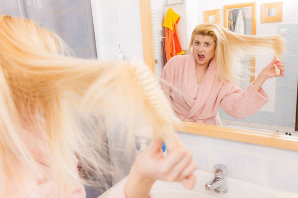 Женщина в халате пытается расчесать свою длинную блондинку очень запутанные волосы, утренняя рутина красоты. Концепция ухода за волосами и укладки
. - Фото, изображение
