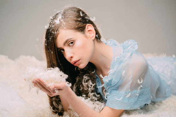 Woman with long hair holds feathers in hand and blow. Girl on calm face lay on bed covered with feathers and fluff. Feather allergy concept. Lady in transparent blue nightie lay on bed. - Photo, image