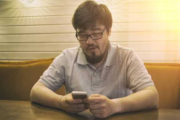 Young Beard Man in Glasses and Grey Shirt sitting in bar looking to the phone and surprised incredible admired. Glad guy with smartphone in right hand - Foto, immagini