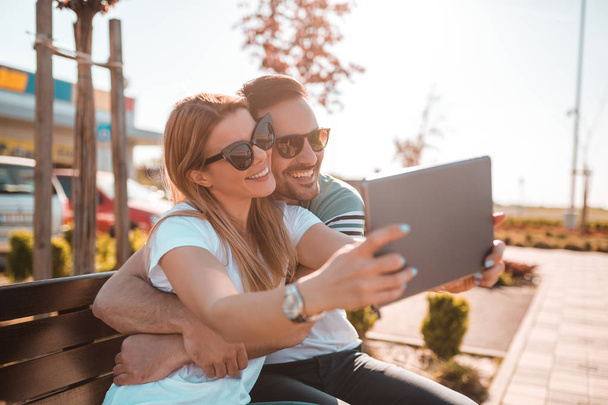 Attractive blonde woman taking selfie with her boyfriend while sitting on a bench and having fun on sunny day - Photo, Image
