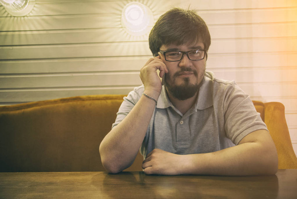 Young Beard Man in Glasses and Grey Shirt sitting in bar talking to the phone and looking at you. Glad guy with smartphone in right hand - Photo, image