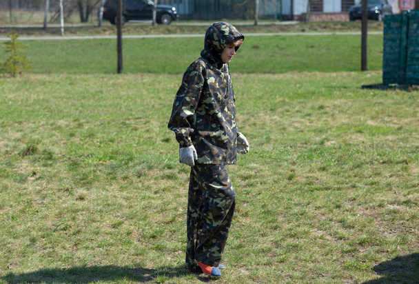 Menino na camuflagem segura uma arma de paintball em uma mão e capacete protetor, de pé no campo com grupo de jogadores no fundo
 - Foto, Imagem
