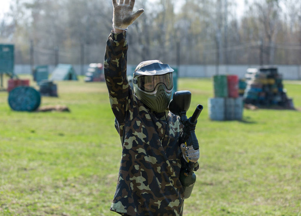 Garçon dans le camouflage tient un pistolet de paintball dans une main et casque de protection, debout sur le terrain avec un groupe de joueurs sur le fond
 - Photo, image