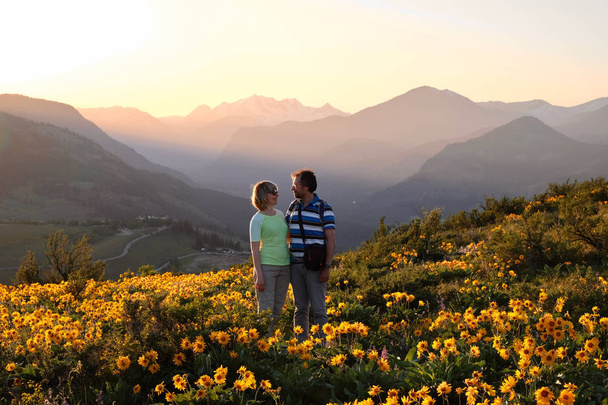 Happy friends surrounded by blooms. Smiling man and woman looking at each other on arnica meadow in bloom. Travel Pacific Northwest. Washington State. United States of America - Photo, Image