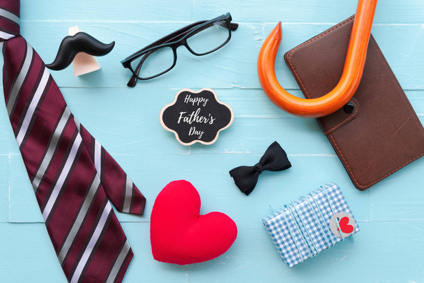 Happy fathers day concept. Red tie, glasses, mustache, notebook, gift box with Happy father's day text and handmade red heart on bright blue pastel wooden table background. - Photo, Image