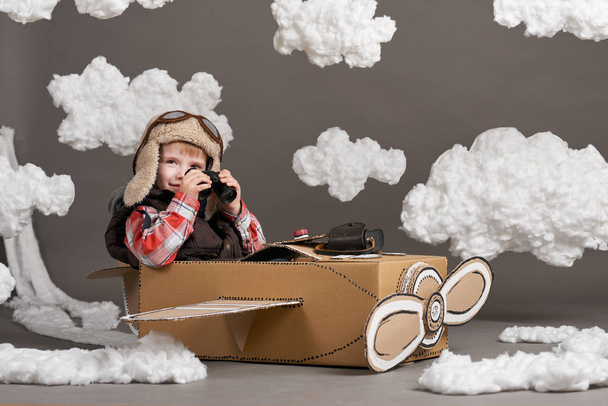 o menino brinca em um avião feito de caixa de papelão e sonha em se tornar um piloto, nuvens de lã de algodão em um fundo cinza
 - Foto, Imagem
