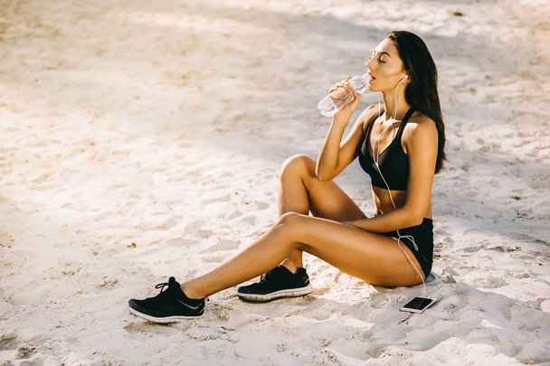 Young pretty sporty style woman sitting on a beach and drinking water - Photo, Image