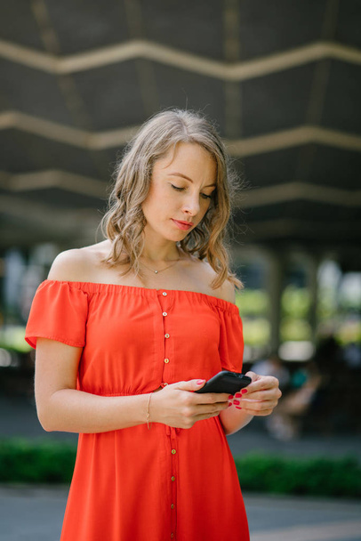 Portrait of a young, attractive Caucasian woman in Asia. She is speaking on her phone in a park and is dressed comfortably in an orange summer dress. - Foto, Imagem