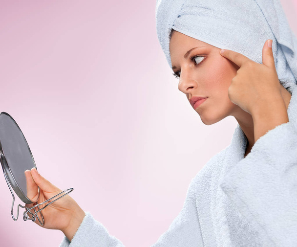 Young woman in bathrobe holding mirror and looking at her skin - Photo, Image