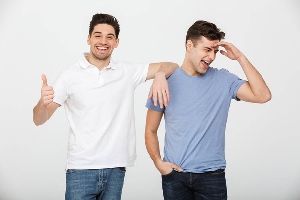 Two handsome men pals 30s wearing casual t-shirt and jeans smiling and posing together on camera with smile thumb up isolated over white background - Foto, Bild