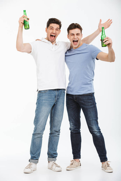 Full length portrait of two happy young men celebrating while holding beer bottles and looking at camera isolated over white background - Photo, Image