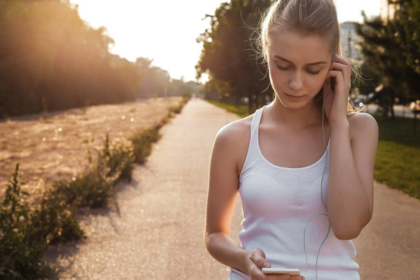 The girl walking outdoor on the road and listen to music in earphones. Woman with blonde hair and in sport clothes looking on her smartphone - Photo, Image