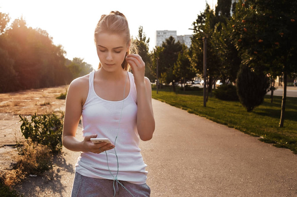 Pretty young girl walking outdoor on the road and listen to music in earphones. Woman with blonde hair and in sport clothes looking on her smartphone - Fotó, kép