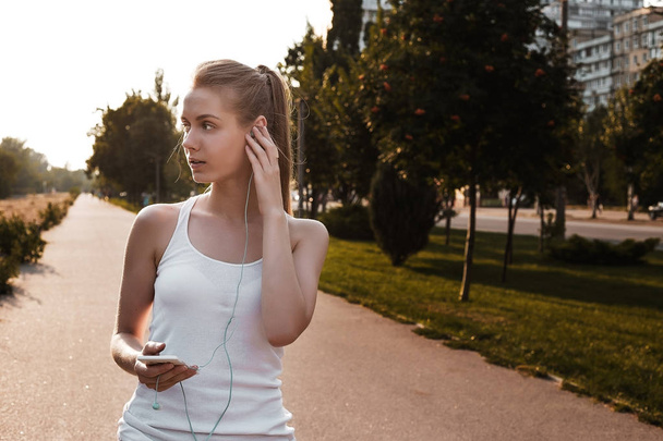 Pretty young girl walking outdoor on the road and listen to music in earphones. Woman with blonde hair and in sport clothes looking sideways - Фото, зображення