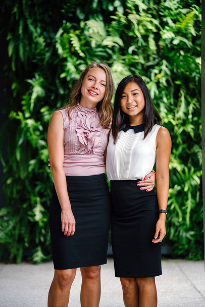 Portrait of a diverse team of two coworkers (a Chinese Asian woman and a Caucasian woman) standing next to one another against a green wall of plants. They are smiling  in a relaxed way. - Foto, imagen