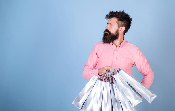 Sale and discount concept. Hipster on serious face carries shopping bags. Man with beard and mustache works as shop assistant, light blue background. Guy shopping on sales season with discounts - Photo, Image