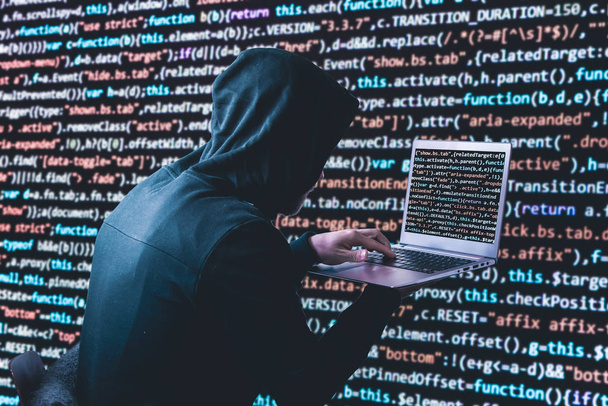 Anonym hacker in a black hoody with laptop in front of a code background with binary streams and information security terms cybersecurity concept - Photo, Image