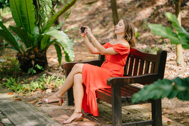 Retrato de uma jovem e atraente mulher caucasiana na Ásia. Ela está falando em seu telefone em um parque e está vestida confortavelmente em um vestido de verão laranja
. - Foto, Imagem