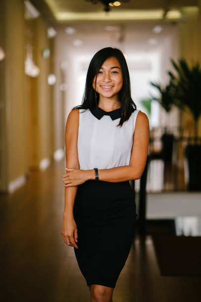 Professional portrait of a young Chinese Asian woman. She is tanned, athletic, attractive and is smiling as she leans against a pillar in the daylight indoor. - Φωτογραφία, εικόνα
