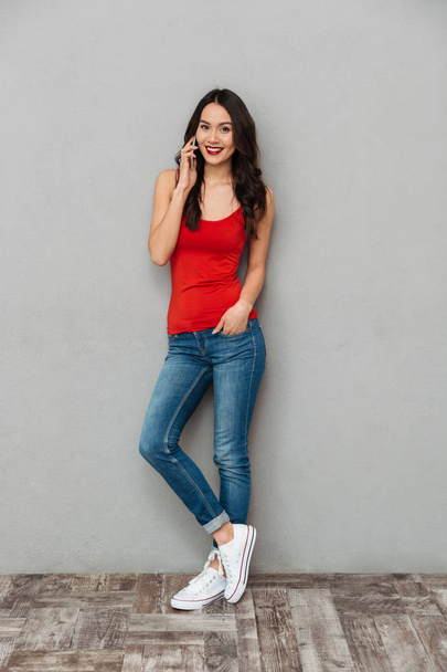 Full length image of Happy brunette woman in casual clothes talking by smartphone while looking at the camera with arm in pocket over grey background - Fotografie, Obrázek