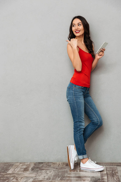 Full length image of Happy brunette woman in casual clothes holding smartphone and pointing back over black background - Foto, afbeelding