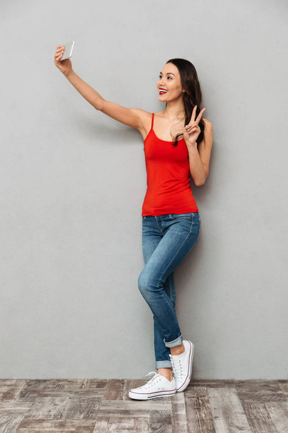 Full length image of Smiling brunette woman in casual clothes making selfie on smartphone while showing peace gesture over grey background - Foto, Imagem