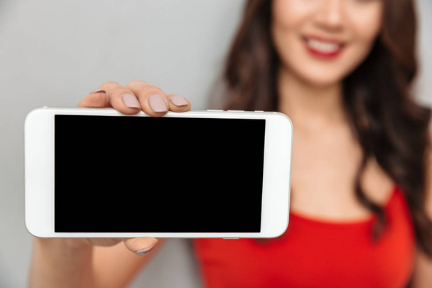 Cropped image of Smiling brunette woman in casual clothes showing blank smartphone screen at camera over grey background. Focus on phone - Foto, Imagen