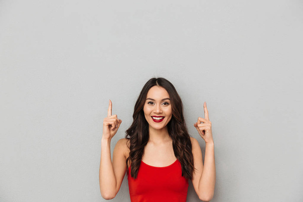 Smiling brunette woman in casual clothes pointing up and looking at the camera over grey background - Fotoğraf, Görsel