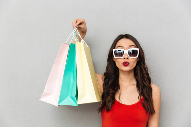 Young brunette woman in casual clothes and sunglasses holding packages and looking at the camera over grey background - Foto, Imagem