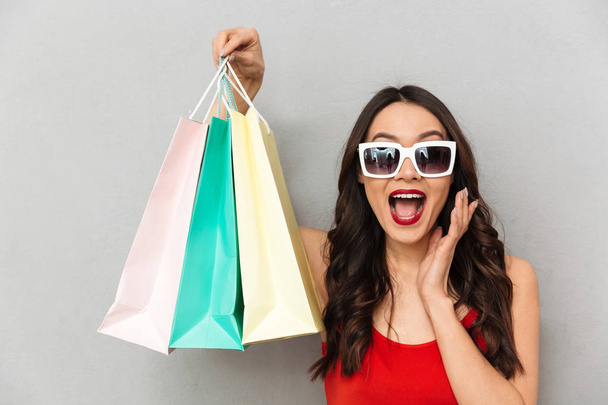 Happy brunette woman in casual clothes and sunglasses holding packages and looking at the camera open mouth over grey background - Foto, Bild