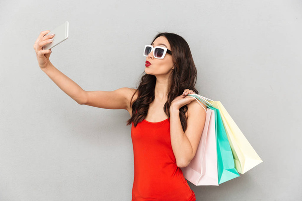 Playful brunette woman in casual clothes and sunglasses making selfie on smartphone while holding packages over grey background - Фото, зображення