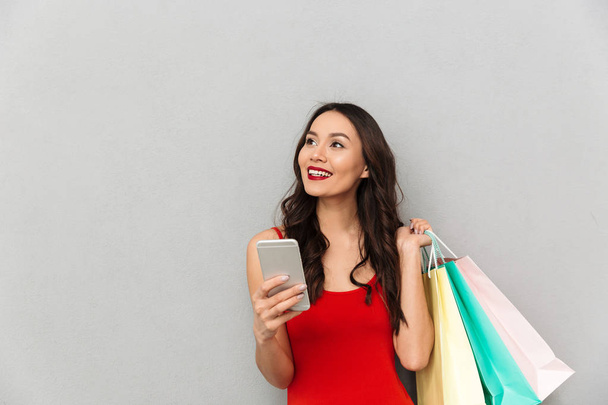 Smiling brunette woman in casual clothes with packages holding smartphone and looking away over grey background - Foto, Imagem