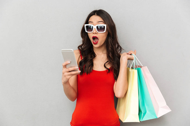 Shocked brunette woman in casual clothes and sunglasses with packages using smartphone over grey background - Φωτογραφία, εικόνα