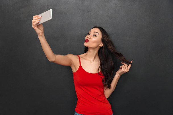 Image of young asian woman standing isolated over dark grey wall background looking aside make selfie by smartphone. - Photo, image