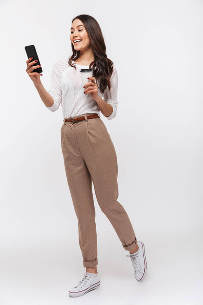 Full length portrait of a smiling asian businesswoman using mobile phone while holding cup of coffee to go isolated over white background - Foto, afbeelding