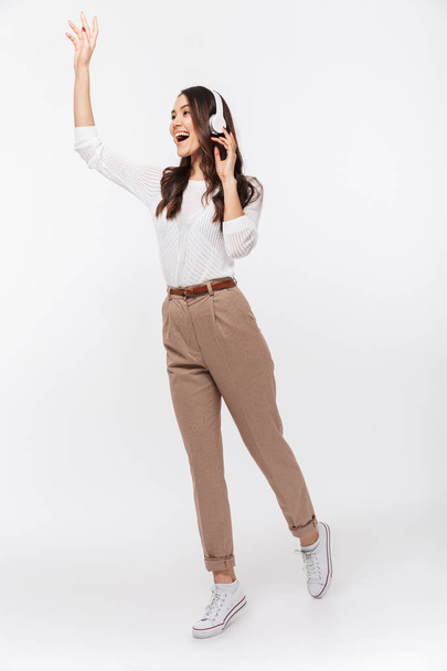 Full length portrait of a joyful asian businesswoman listening to music with headphones while waving isolated over white background - Фото, зображення