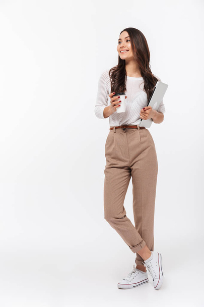 Full length portrait of a satisfied asian businesswoman carrying laptop computer and cup of coffee to go while standing isolated over white background - Fotoğraf, Görsel