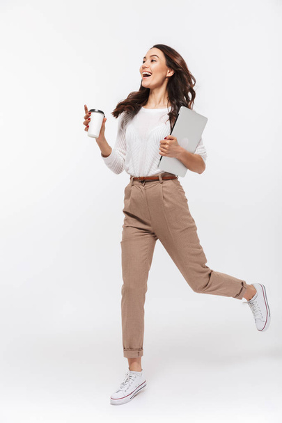Full length portrait of a happy asian businesswoman carrying laptop computer and cup of coffee to go while running isolated over white background - Photo, image