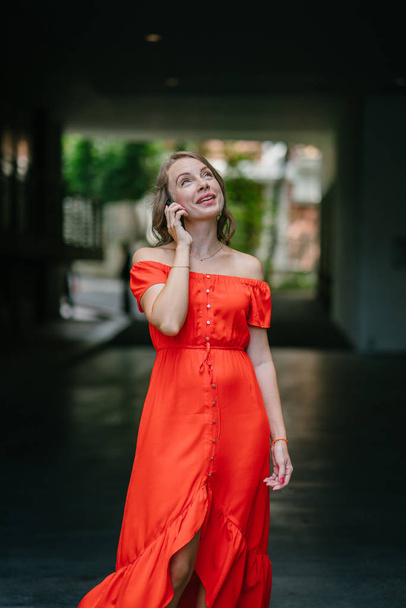 Portrait of a young, attractive Caucasian woman in Asia. She is speaking on her phone in a park and is dressed comfortably in an orange summer dress. - Foto, Imagen