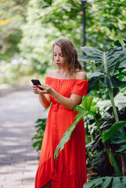 Portrait of a young, attractive Caucasian woman in Asia. She is speaking on her phone in a park and is dressed comfortably in an orange summer dress. - Photo, Image
